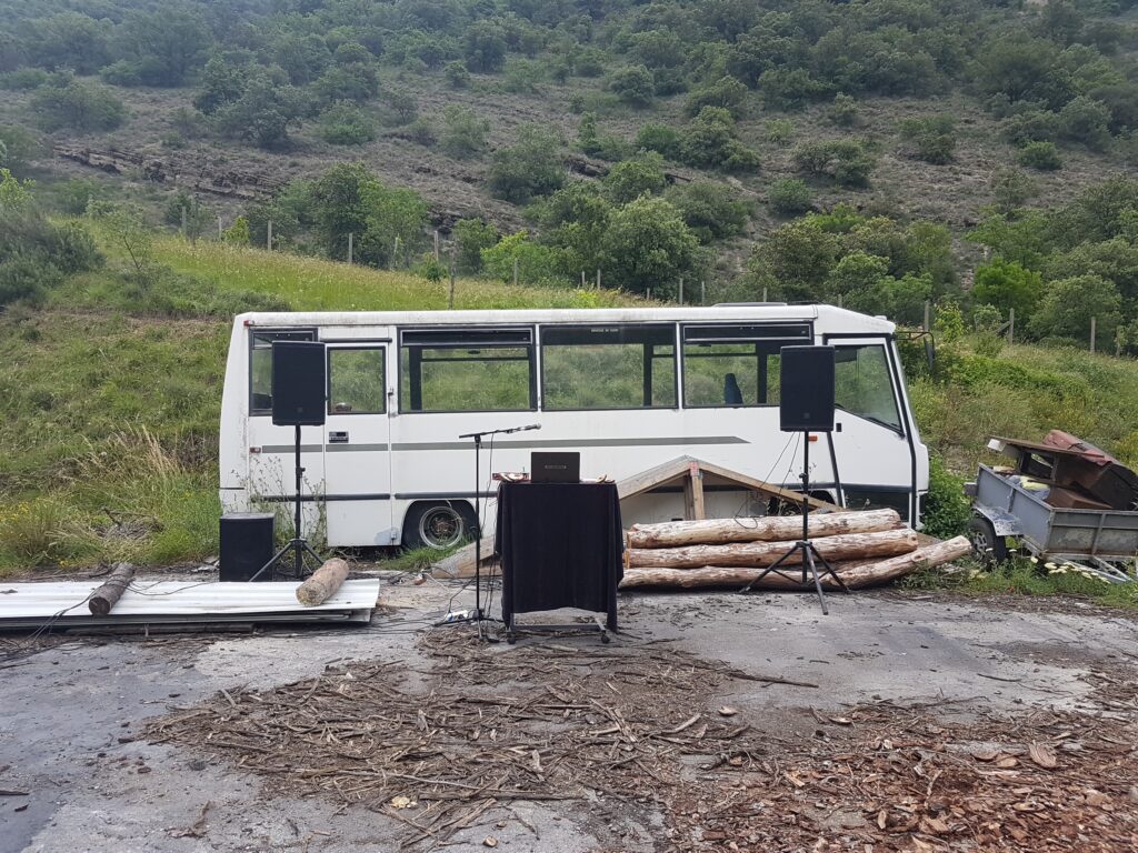 Un bus abandonné git devant une montagne, devant lui sont entreposés de gros troncs d'arbres débités, deux grosses enceintes de diffusions et un petit pupitre sur lequel trône mon ordinateur.