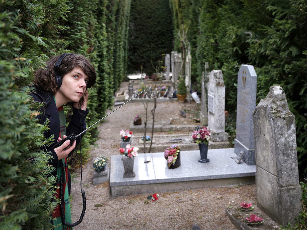 Samia écoute la radio dans un cimetière en passant sa tête entre deux murs de cyprès. Elle tient son casque comme si elle écoutait quelque chose.