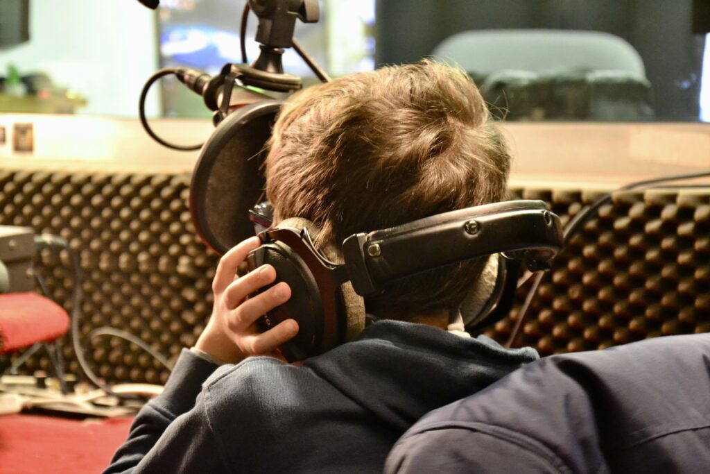 Un enfant serre un casque trop grand pour lui contre ses oreilles. Il a l'air de parler dans le micro d'un studio radio.