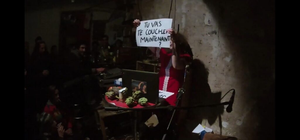 Samia dans une sublime robe rouge lève un petit panonceau sur lequel est écrit "Tu vas te coucher maintenant ?" Elle est visiblement dans une salle de concert, derrière son ordinateur et quelques artichauts, en train de performer,