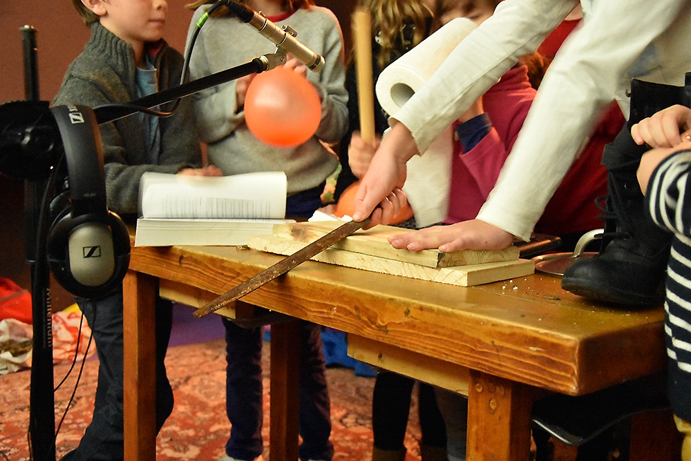 Plusieurs enfants sont réunis autour d'une table vers laquelle pointe un micro. Une main frotte une grosse queue de rat contre des planches de bois. Un autre enfant tient un ballon de baudruche.