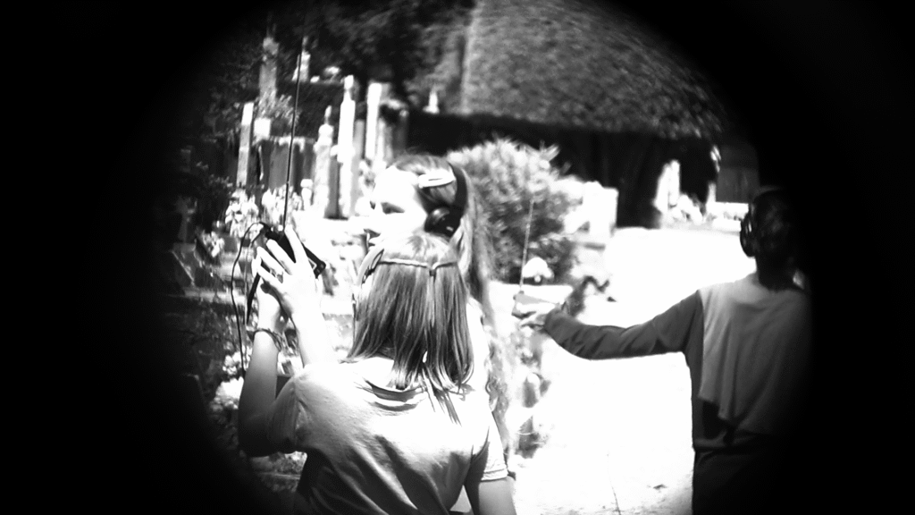 Deux adolescentes dans un cimetière lèvent une radio vers le ciel. Une femme à l'arrière lève elle aussi sa radio. Tout le monde a un casque sur les oreilles.